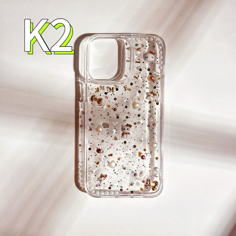 May include: Clear phone case with a glittery, liquid-like design. The case has small, colorful charms and glitter inside. The case has the text "AMME" printed on the back.