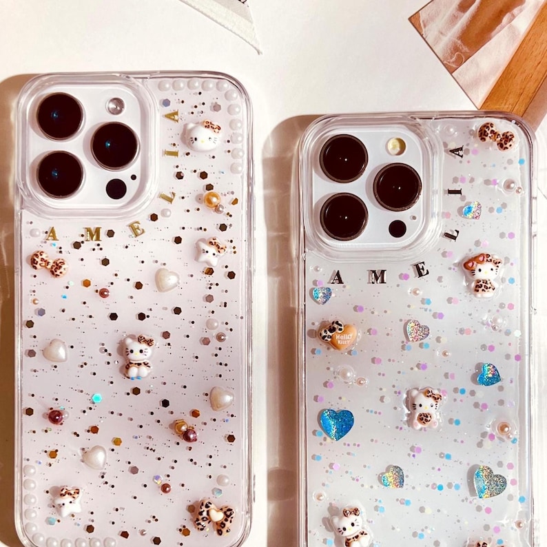 May include: Two clear phone cases with glitter and small charms. The cases have the name "Amelia" written on them. One case has a pink background and the other has a white background.