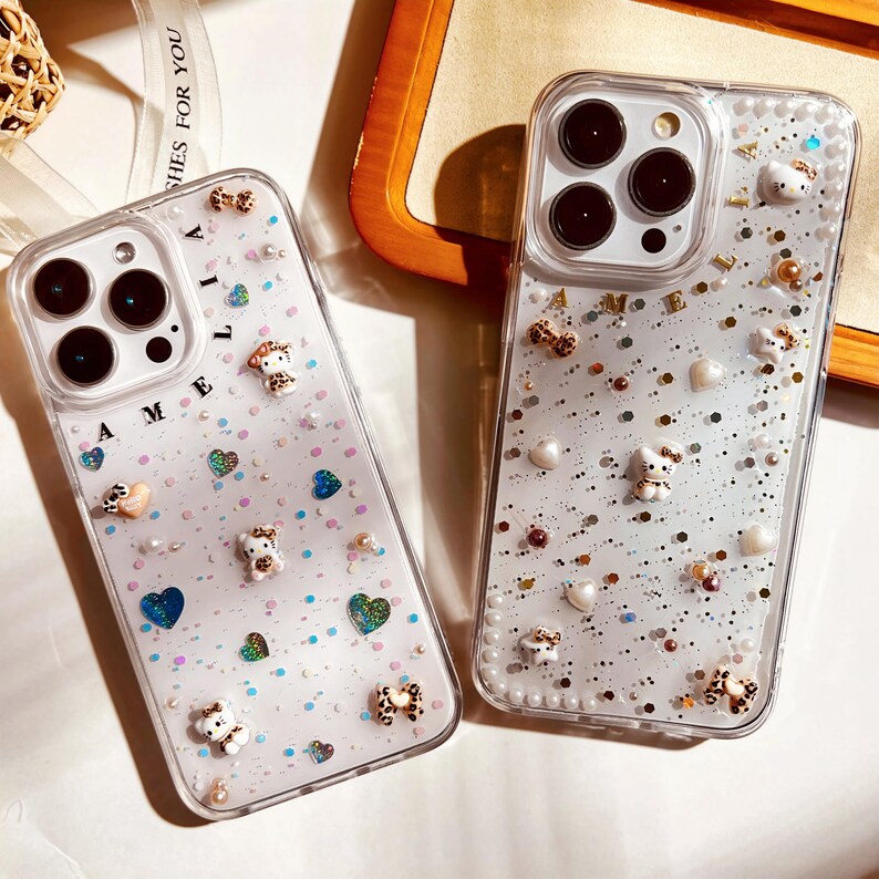 May include: Two clear phone cases with glitter and small decorative charms. The cases have the word "Amelia" written on them.