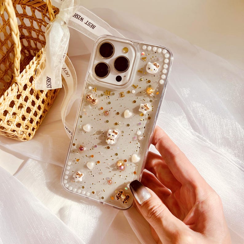Puede incluir: Una funda transparente para tel&eacute;fono con un fondo brillante y peque&ntilde;as figuritas de animales blancas. La funda tiene un borde elevado de peque&ntilde;as perlas blancas.