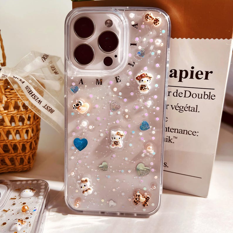 May include: A clear phone case with a glittery background and a variety of small charms, including hearts, stars, and cartoon animals. The case has the name "Amelia" written on the back.