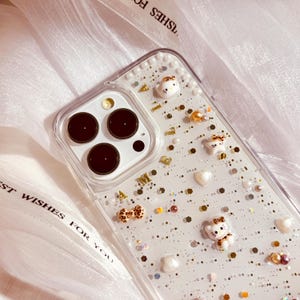 Puede incluir: Una funda transparente para tel&eacute;fono con fondo blanco y purpurina dorada. La funda est&aacute; decorada con peque&ntilde;as perlas blancas, letras doradas y peque&ntilde;os osos de dibujos animados. La funda est&aacute; dise&ntilde;ada para un iPhone.