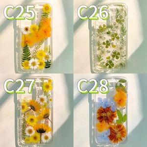 Op de afbeelding: Vier transparante telefoonhoesjes met geperste bloemen. De hoesjes hebben gele, witte en oranje bloemen en groene bladeren. Elk hoesje heeft een ander bloemarrangement, waaronder madeliefjes, zonnebloemen en klavers met vier blaadjes. De hoesjes zijn gelabeld met C25, C26, C27 en C28.