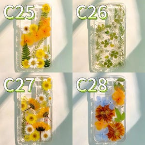 Pode incluir: Quatro capas de telefone transparentes com flores prensadas. Cada capa apresenta um arranjo floral diferente: margaridas amarelas, flores laranja, fetos verdes e flores azuis e laranja. As capas est&atilde;o rotuladas C25, C26, C27 e C28.