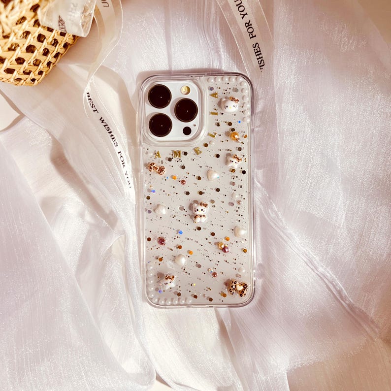Puede incluir: Una funda transparente para tel&eacute;fono con un fondo brillante y peque&ntilde;os osos blancos, perlas y otros adornos. La funda tiene una lente de c&aacute;mara elevada y un borde transparente.