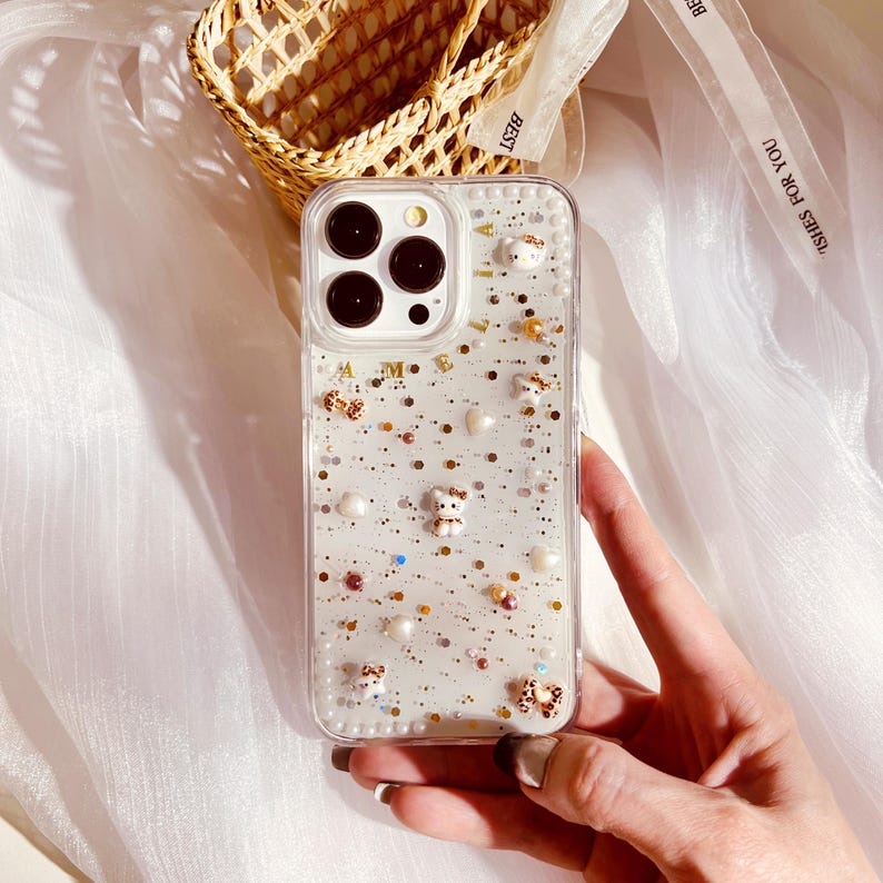 Puede incluir: Una funda transparente para tel&eacute;fono con fondo brillante y peque&ntilde;os osos blancos, lazos y perlas. La funda tiene una inscripci&oacute;n dorada que dice "AMIE".