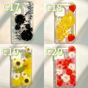 Pode incluir: Capas de telefone transparentes com flores secas e frutas embutidas. Os designs incluem flores pretas, fatias de laranja e morangos, girass&oacute;is amarelos e flores vermelhas e brancas. Cada capa &eacute; rotulada com uma letra e um n&uacute;mero.