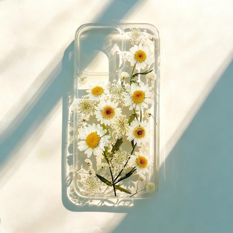 May include: Clear phone case with embedded white daisies and yellow centers. The case has a rectangular shape with rounded corners and cutouts for the camera and buttons. The flowers are arranged in a natural, organic pattern, creating a unique and stylish accessory.