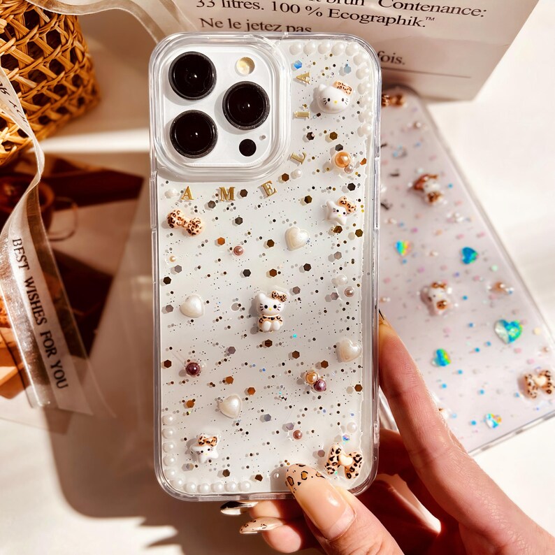 May include: A white iPhone case with a glittery, sparkly design. The case has a clear back and a white border. There are small white and gold charms on the case, including hearts, stars, and a small cat. The case is decorated with the letters "A M E"