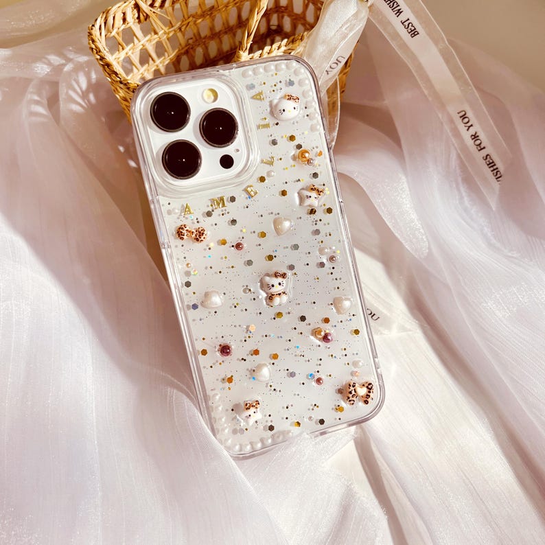 Puede incluir: Una funda transparente para tel&eacute;fono con un fondo brillante y varios adornos, incluyendo peque&ntilde;as perlas blancas, lazos dorados y peque&ntilde;os osos de dibujos animados. La funda est&aacute; dise&ntilde;ada para un iPhone con una lente de c&aacute;mara cuadrada.