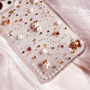 Puede incluir: Una funda transparente para tel&eacute;fono con un fondo brillante y varios encantos, incluyendo corazones, estrellas y peque&ntilde;as figuras de animales. La funda tiene un borde de perlas alrededor del borde.