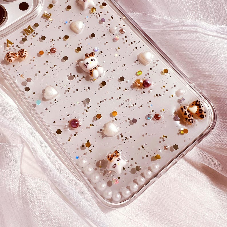 Puede incluir: Una funda transparente para tel&eacute;fono con un fondo brillante y varios encantos, incluyendo corazones, estrellas y peque&ntilde;as figuras de animales. La funda tiene un borde de perlas alrededor del borde.