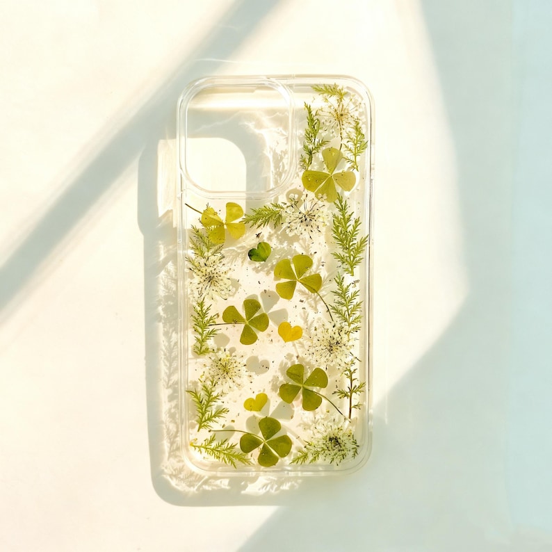 May include: Clear phone case with embedded dried green clovers, small yellow hearts, and white flowers. The case has a rectangular shape with rounded corners and a transparent design, showcasing the natural elements.