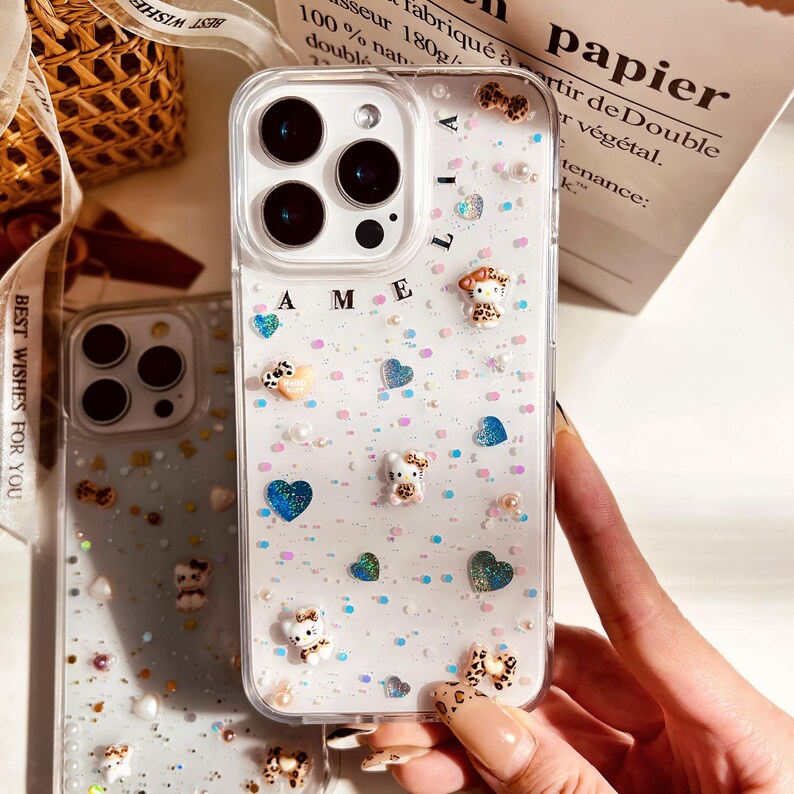 May include: A clear phone case with a white background and glitter. The case has a variety of small, colorful decorations, including hearts, stars, and small animal figurines. The case also has the letters "AME" printed on it.