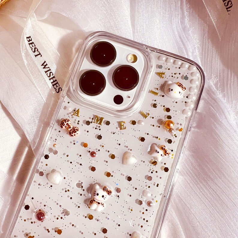 Puede incluir: Una funda transparente para tel&eacute;fono con fondo de purpurina blanca y letras doradas que dicen "AMELLA". La funda tiene una variedad de peque&ntilde;as decoraciones blancas, como perlas, y peque&ntilde;as figuras de gatos de dibujos animados blancas.