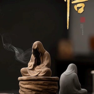 May include: A serene, brown ceramic incense burner featuring a meditating figure with hands clasped in prayer. Smoke gently rises from the burner, creating a peaceful ambiance. A second figure sits nearby. The word "悟" is written in gold.