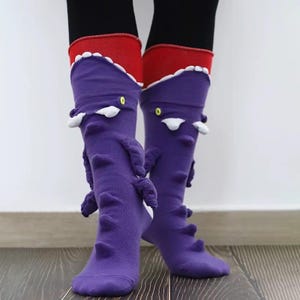 May include: Purple knee-high socks with a monster design. The socks have white teeth, yellow eyes, and textured details. The top of the socks are red with a white scalloped edge. The socks are worn over black leggings.