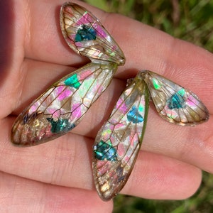 May include: A pair of iridescent butterfly wings with a pink, green, and gold color scheme. The wings are made of resin and have a delicate, translucent appearance.