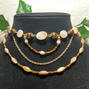 Puede incluir: Un collar de varias capas con cadenas doradas, cuentas marrones de ojo de tigre y cuentas blancas de concha.