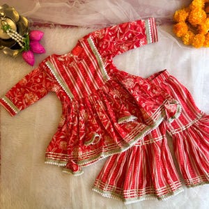 May include: A red and white striped and floral print girls' outfit with white lace trim. The top is a short-sleeved, ruffled, and flared style. The bottom is a pair of flared pants with a bow detail.