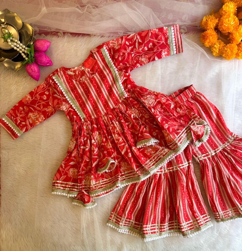 May include: A red and white striped and floral print girls' outfit with white lace trim. The top is a short-sleeved, ruffled, and flared style. The bottom is a pair of flared pants with a bow detail.