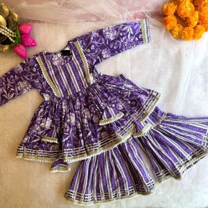 May include: Purple and white floral print girls' dress with a layered skirt and gold trim. The dress has a long sleeve and a round neckline.
