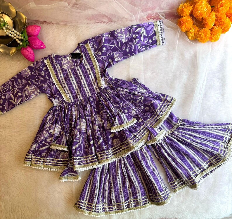 May include: Purple and white floral print girls' dress with a layered skirt and gold trim. The dress has a long sleeve and a round neckline.