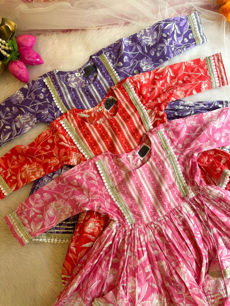 May include: Three girls' dresses in pink, red, and purple with floral patterns and white lace trim. The dresses have a striped bodice and a gathered skirt.