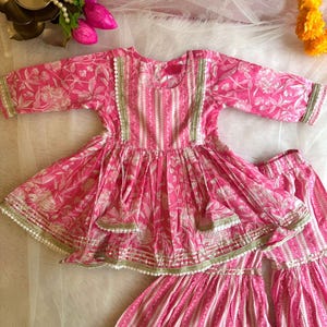 May include: A pink and white floral print girls' dress with a layered skirt and matching pants. The dress has a round neckline and three-quarter sleeves. The outfit is decorated with gold and white trim.