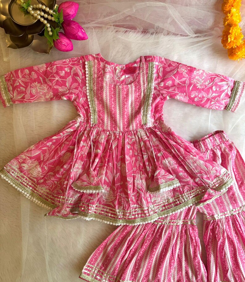 May include: A pink and white floral print girls' dress with a layered skirt and matching pants. The dress has a round neckline and three-quarter sleeves. The outfit is decorated with gold and white trim.