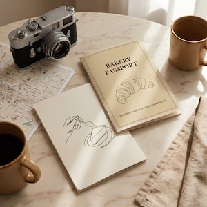 Może przedstawiać: Beżowa broszura zatytułowana BAKERY PASSPORT z ilustracją croissanta i tekstem Proof that carbs are a solid personality trait, obok białego notatnika z rysunkiem liniowym dłoni trzymającej łyżkę nad kawałkiem ciasta.