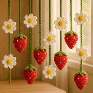 Könnte beinhalten: Gehäkelte Erdbeer- und Gänseblümchen-Girlande. Die Girlande zeigt rote Erdbeeren mit grünen Spitzen und weiße Gänseblümchen mit gelben Zentren, auf grüner Schnur aufgereiht. Geeignet als Frühlings- oder Sommerdekoration.