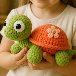 Puede incluir: Una tortuga de ganchillo hecha a mano en tonos verdes y naranjas. La tortuga tiene ojos grandes y un detalle de flor en su caparazón. El juguete es suave y parece estar hecho de hilo.