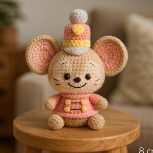 Puede incluir: Una muñeca de ratón de ganchillo, de aproximadamente 8 cm de altura, está sentada en un taburete de madera. El ratón tiene orejas grandes, un atuendo rosa y un sombrero con detalles amarillos y rosas. La muñeca tiene una cara sonriente y pequeños bigotes.