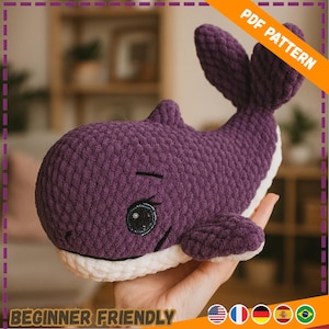 Puede incluir: Una ballena de ganchillo morada hecha a mano con detalles blancos, con ojos bordados y una expresión sonriente. La imagen incluye el texto "PDF PATTERN" y "BEGINNER FRIENDLY".