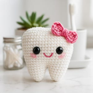 Może przedstawiać: Szydełkowy biały ząb z różową kokardą, czarnymi oczami i uśmiechniętymi ustami. Ząb ma różowe rumieńce na policzkach. Tło obejmuje małą roślinę sukulentową w białej doniczce, słoik i szczoteczkę do zębów.