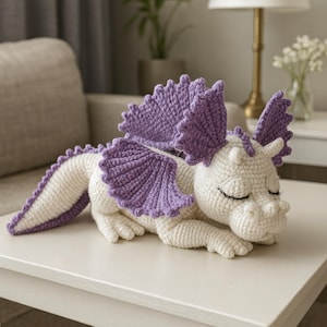 May include: A handmade crochet dragon in white and purple. The dragon has closed eyes, a long tail, and large, textured wings. The dragon is lying down on a white surface.