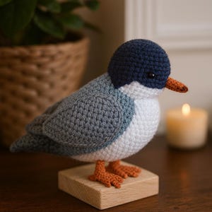 Puede incluir: Figura de pájaro de ganchillo hecha a mano. El pájaro presenta una cabeza azul marino, alas grises y un pecho blanco. Tiene un pico y patas naranjas, y está montado sobre un pequeño bloque de madera. El fondo incluye una vela encendida y una cesta tejida.