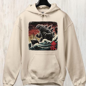 May include: Sand-coloured hoodie with a Godzilla graphic in a vintage Japanese art style. The design shows Godzilla emerging from ocean waves, attacking a ship. The graphic is in a square frame with Japanese text.