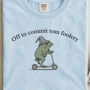 Peut inclure: T-shirt bleu clair avec l'inscription "Off to commit tom foolery" au-dessus d'une grenouille de dessin animé portant un chapeau de sorcier et conduisant un scooter. Le t-shirt a un col rond et une étiquette en haut.