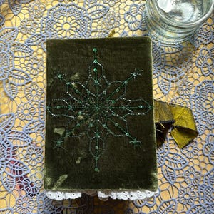 May include: A dark green velvet journal with a snowflake design embroidered in green sequins. The journal has a white lace trim and a gold ribbon closure. A clear glass jar with a lid is in the background.