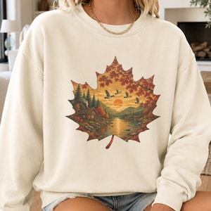 Op de afbeelding: Een crèmekleurige sweater met ronde hals en een grote afbeelding van een esdoornblad op de borst. Het blad bevat een gedetailleerde landschapsillustratie van een hut aan een meer, dennenbomen, vliegende eenden en een zonsondergang.