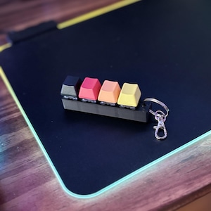 May include: A keychain with a black, red, orange, and yellow keycap holder. The keycaps are attached to a silver metal keychain.
