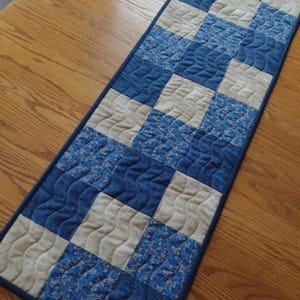May include: A quilted table runner featuring a patchwork design in shades of blue and beige. The runner has a dark blue border and a wavy quilted pattern. The squares alternate between solid blue, patterned blue, and beige.