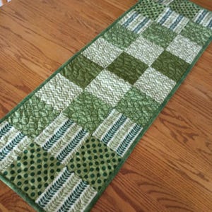 May include: A rectangular quilted table runner featuring a patchwork design of alternating green fabric squares. The patterns include dark green polka dots, light green leaf prints, wavy lines, and geometric circles, all finished with a green border.