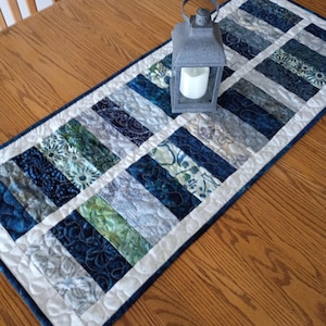May include: A rectangular quilted table runner featuring a patchwork design of blue, green, and white fabric strips with various floral and abstract patterns. A gray metal lantern with a white pillar candle sits on top of the runner.
