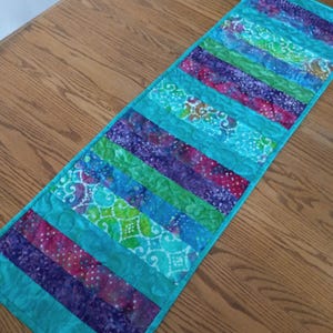 May include: A colorful quilted table runner with a vibrant design. The runner features alternating stripes of turquoise, purple, green, and pink fabrics. The fabrics have patterned designs. The edges are finished with a turquoise border.