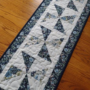 May include: A quilted table runner with a floral pattern. The runner features a white center with dark blue and floral patterned triangles. The border is dark blue with a floral design. The runner is approximately 36 inches long.