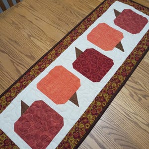 May include: A rectangular quilted table runner featuring a white center panel with five alternating orange and dark red pumpkin shapes, each with a brown stem. The runner is bordered by a dark brown fabric with a small yellow and red floral pattern.