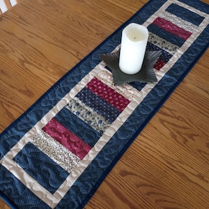 May include: A rectangular quilted table runner featuring a patchwork design of navy blue, cream, and burgundy fabric strips with floral patterns. A dark gray star-shaped candle holder sits on the runner, holding a white pillar candle.